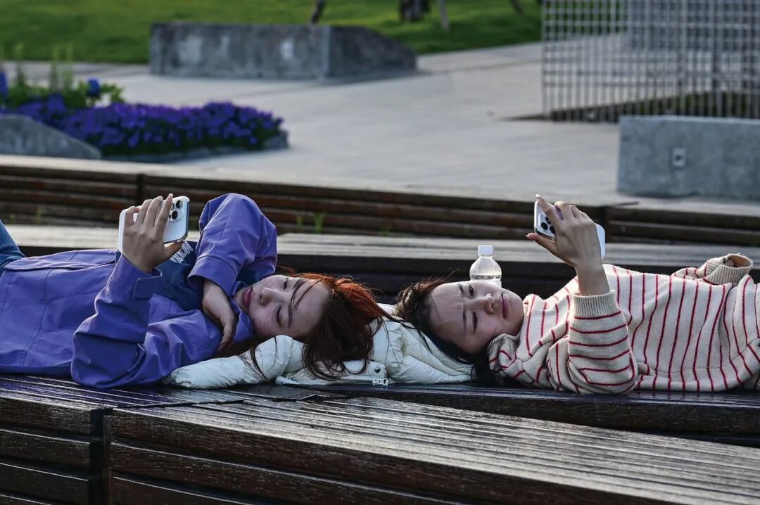 2026 年 4 月 9 日,上海。两名女性躺在黄浦江畔的长椅上看手机。(图 /AFP/CFP)