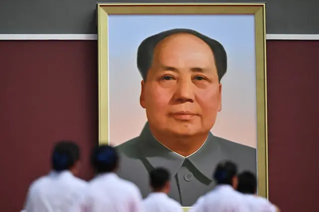 一幅大型毛泽东肖像悬挂在红色墙壁上，照片从下方拍摄，前景有数名身穿浅色衣服的人站立，面向肖像。