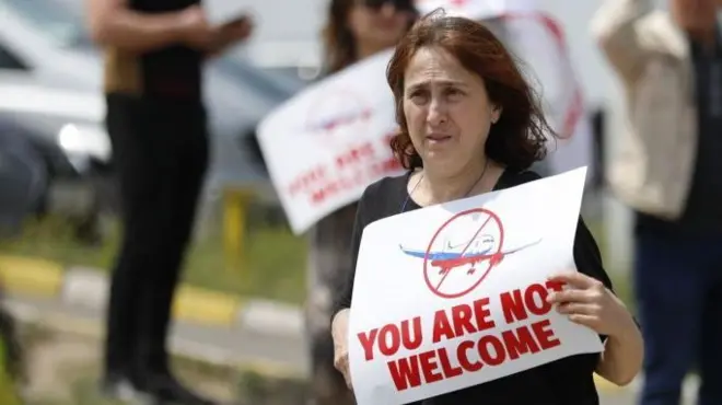 Женщина с плакатом You are not welcome и изображением перечеркнутого самолета в цветах российского флага