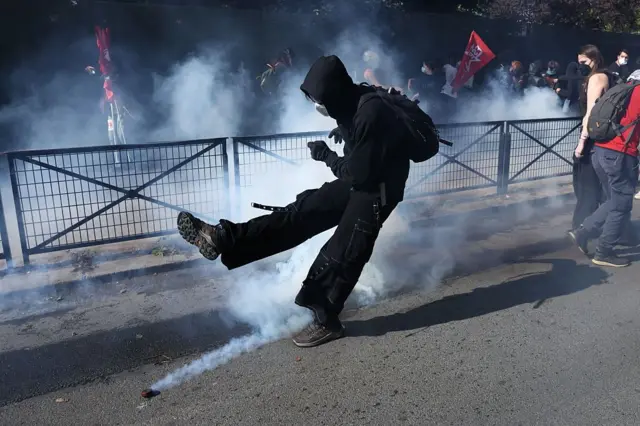 Школьные протесты во Франции: молодой человек во всем черном, в капющоне, маске и рюкзаком за плечами поддает ногой дымящуюся пиротехнику