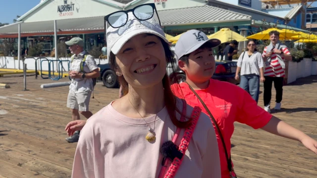 浅井舞子站在画面中央，戴着一顶棒球帽，帽簷上方放着一副眼镜。她的儿子站在她身后，穿着红色上衣。他们站在一条木制步道上，背景是一家海鲜餐厅。