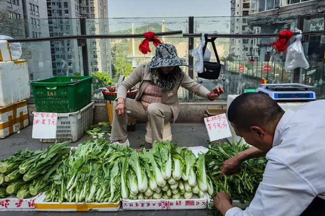 2025年4月17日，中国重庆，两名郊区农民在高架人行道上的非正式摊位上贩售新鲜采摘的蔬菜，男子正在整理农产品，女子坐在他身后。