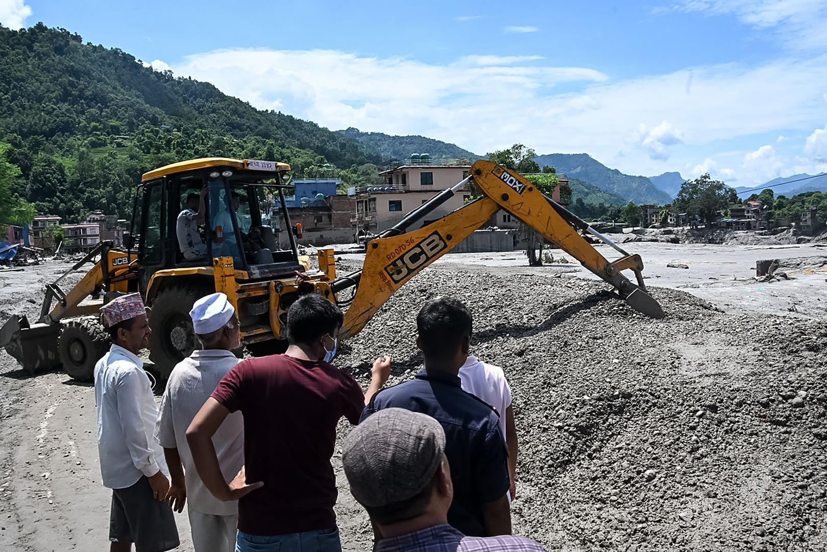 ▲当地时间2026年8月27日，尼泊尔努瓦果德县德维加特地区，居民聚集在道路旁，工人使用推土机清理山洪暴发后的泥浆和碎石（图据视觉中国）