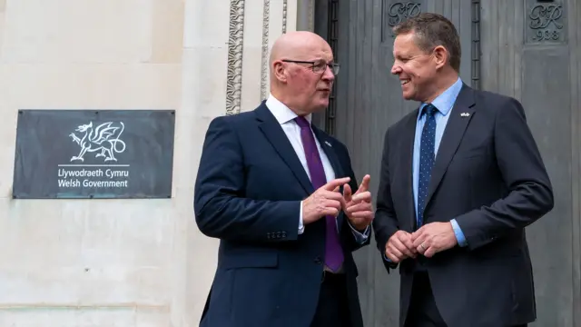 约翰·斯文尼（John Swinney）、杨伟思（Rhun ap Iorwerth）站在威尔士政府总部的一扇金属门外，从照片的左侧可以看到一块铭牌。