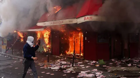 На снимке, сделанном Getty Images, мужчина снимает на видео, как из бара «La Barrique» поднимается дым во время демонстрации против «высокой стоимости жизни» в Париже.