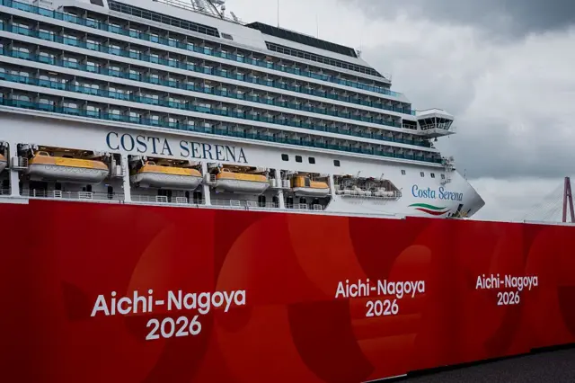 意大利邮轮Costa Serena停泊于阴云密布的天空下,前景可见写有“Aichi-Nagoya 2026”的大型标志。