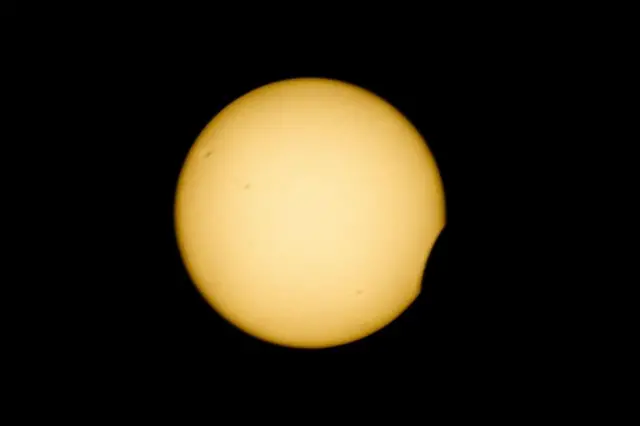 一轮黄色圆盘状的太阳悬挂在漆黑天空中。随着月球从其前方移过,太阳右下角出现一道细小的新月形缺口,仿佛被咬去了一小口。