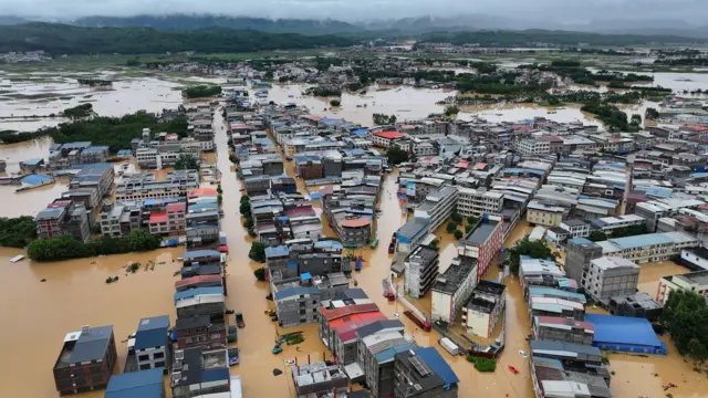 洪水淹没中国南宁横州市的住宅与建筑，一片棕色洪水汪洋之中，背景可见绿色山脉及阴云密布的天空。（中新社图片6/7/2026）