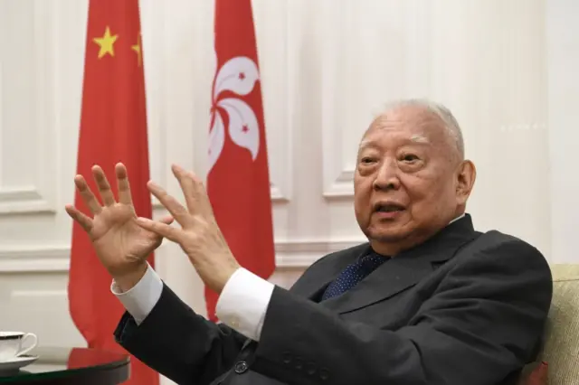 董建华坐在沙发上，背景是中国国旗和香港区旗