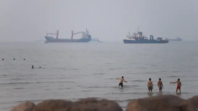 朦胧天色下，近岸停泊着货船及油轮，前景可见有人在沿岸海域游泳及涉水。（30/6/2026）