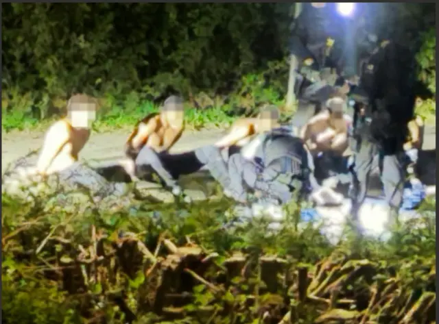 在韦尔福德（Whelford）被拘留的男子。他们的脸部经过了模糊处理。他们赤裸着上身。背景中有成排的树木。（27/9/2026）