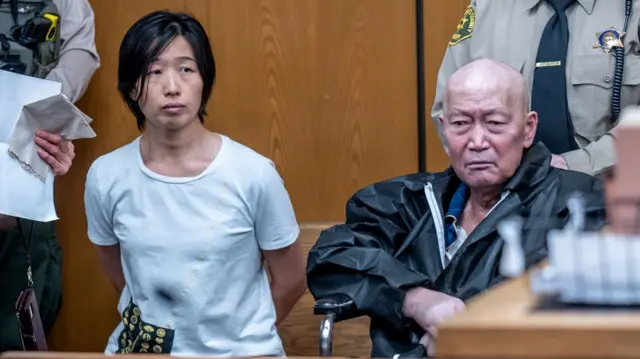 左为张思薇,右为宣国军,两人坐在帕萨迪纳法庭内,身后站着两名警长。张思薇双手背在身后,身穿蓝色衬衫;宣国军身穿黑色夹克。